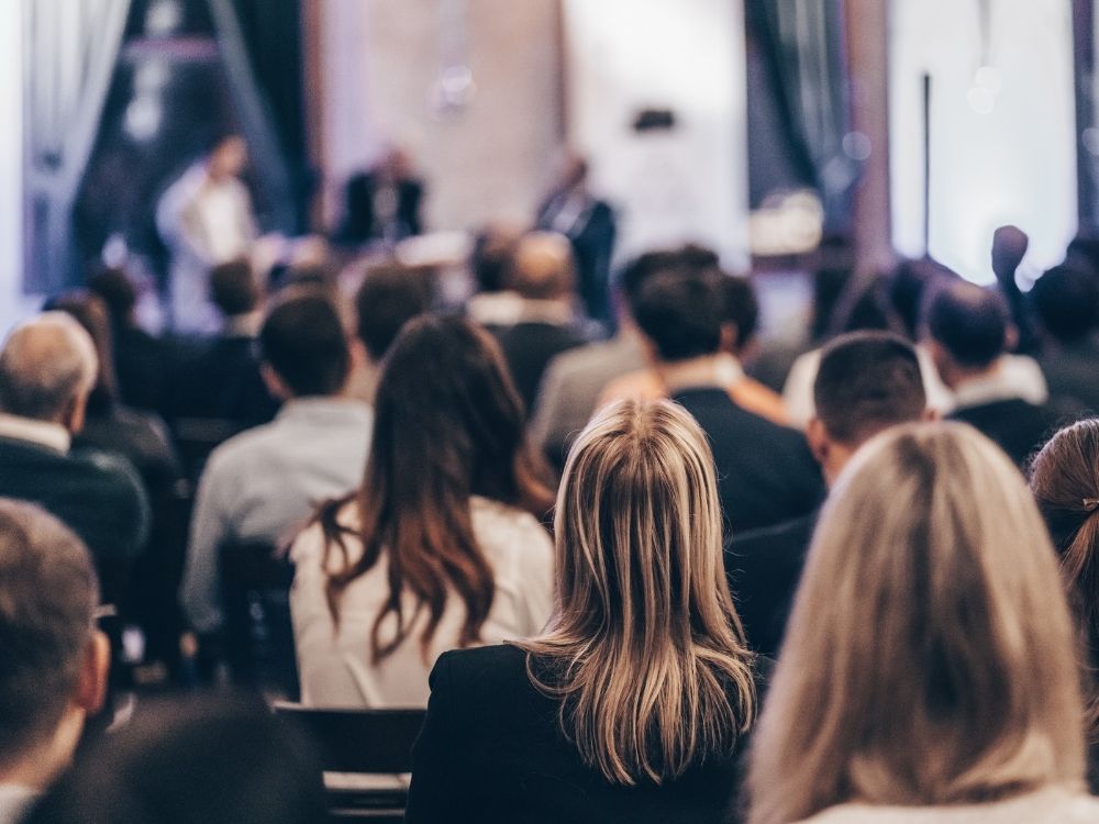 A view from the back of a meeting room; attendees watch a panel present.