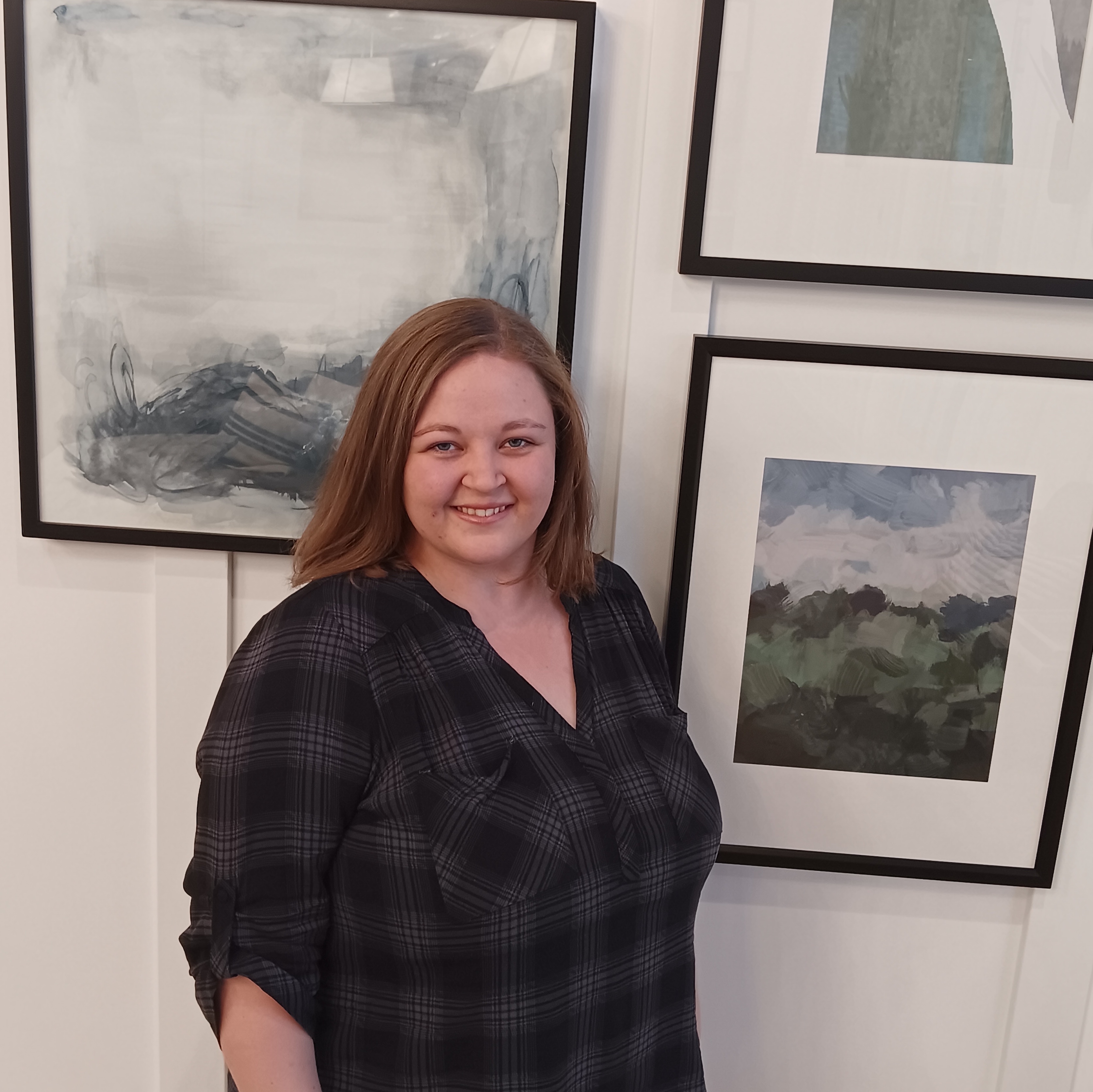 Alyssa is smiling standing in front of a white wall with three paintings hanging behind her.  She has long light brown hair and is wearing a black and gray top.