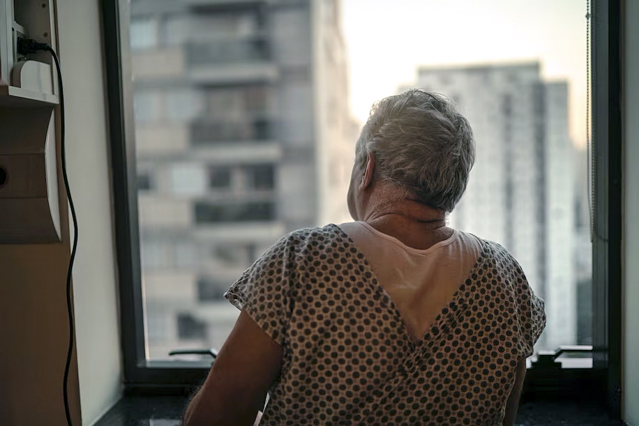 Male senior citizen in hospital gown looking out window Male senior citizen in hospital gown looking out window