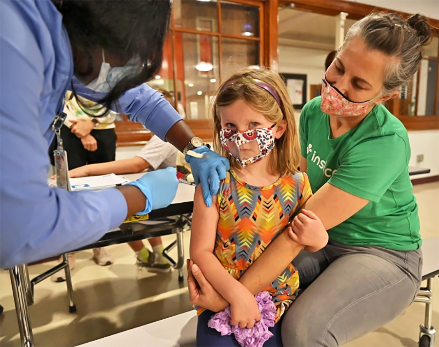 Camden VanBuskirk, 5, gets her first COVID-19 vaccine shot from medical assistant Yaa Prempeh with her mother Monica, at West High School in Denver