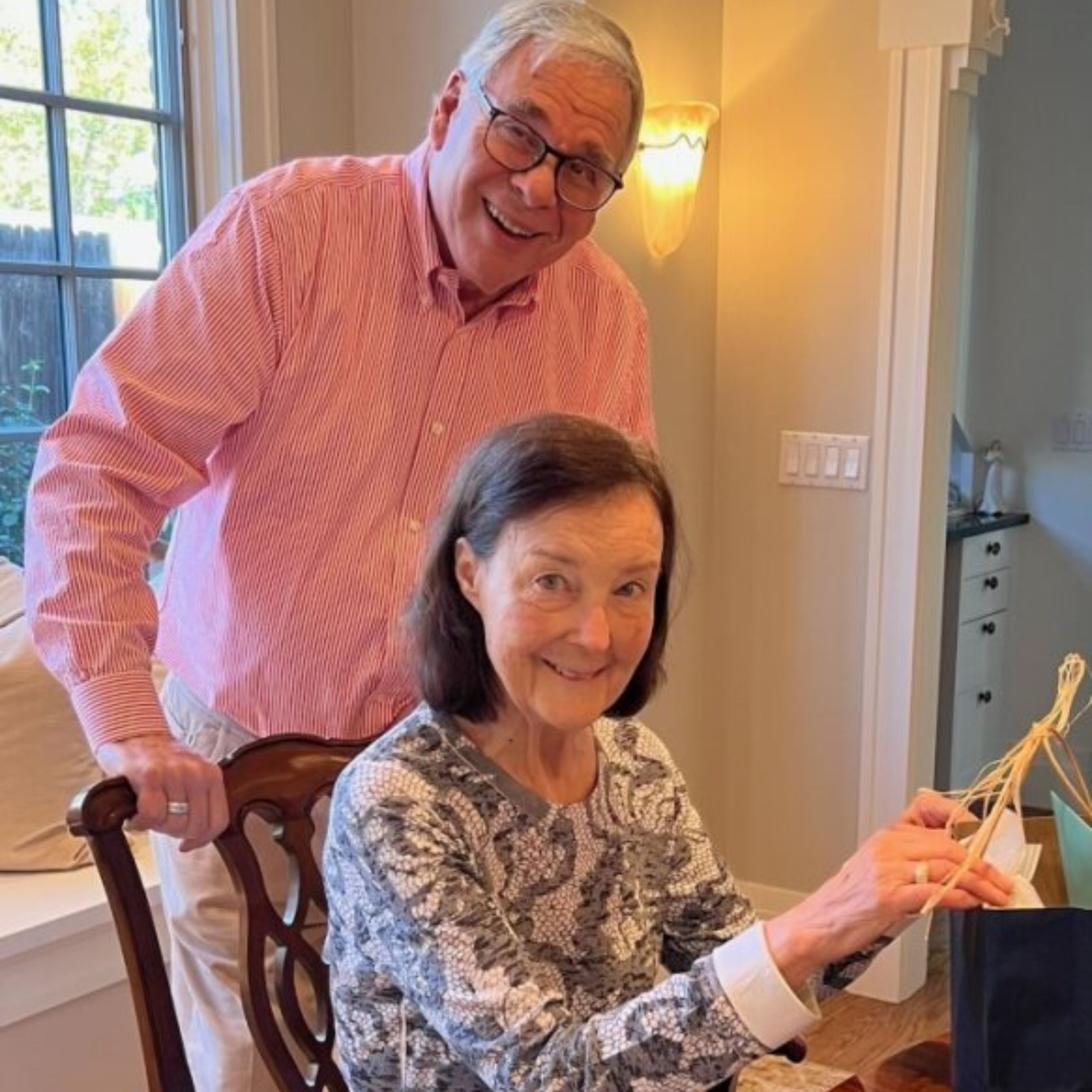 The late Dr. Mary Krugman smiles as she opens a gift while sitting in a chair, her husband, Dr. Dick Krugman, stands behind her.