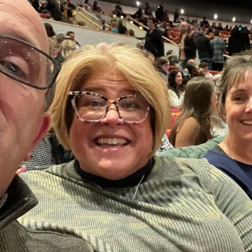SOC Article Feature Image Stacey Huelsing-Dhein Stacey Huelsing-Dhein smiling broadly while sitting in the audience of a sporting stadium with loved ones.