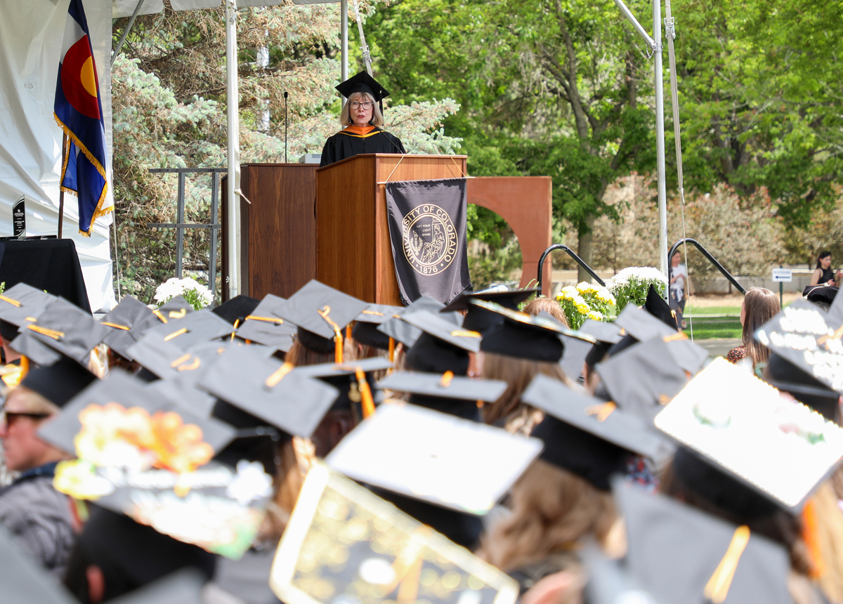 Karen Zink speaks to graduating students from a podium. All are wearing graduation caps and gowns.