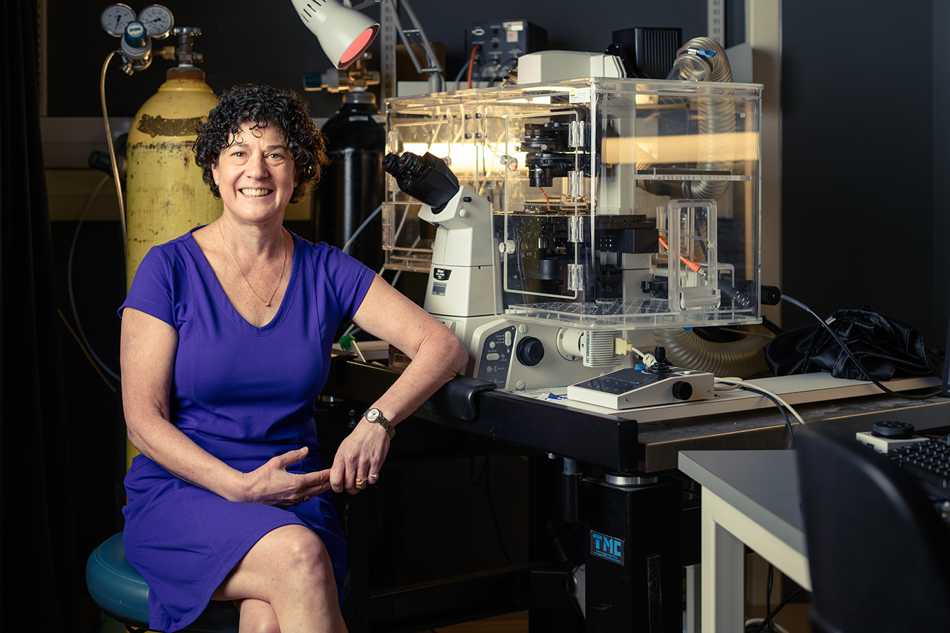 Dr. Sussel sits at a desk in a purple dress, in front of complicated lab equipment and a yellow air canister.