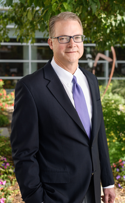 A photo of Dr. Messersmith in a black suit standing in front of green trees and bushes.