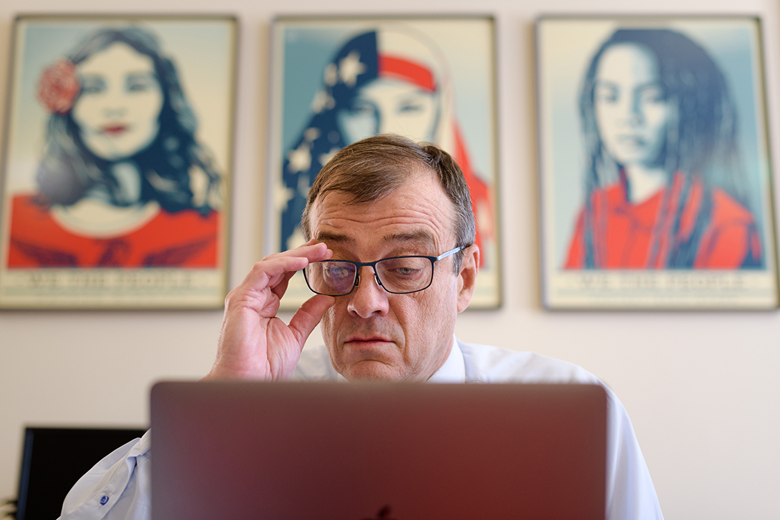 Dean Reilly, sitting at his desk, adjusts his glasses. Behind time, there are three paintings of young women of color.