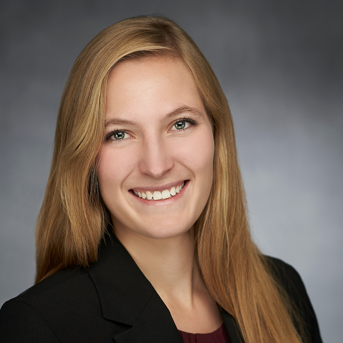 Courtney Olsen, medical scholar. She is wearing a black blazer and is smiling at the camera in front of a light gray photography studio background.