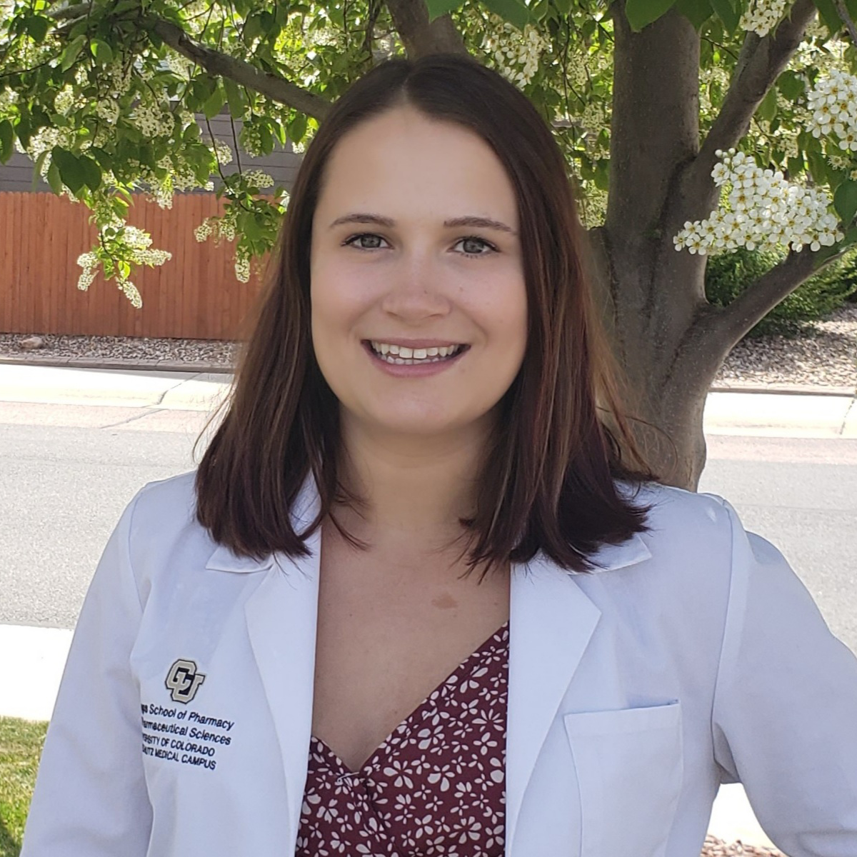 Paige Johnson, smiling at the camera. She is wearing a white pharmacy coat over a maroon shirt and standing in front of a small tree and a street.