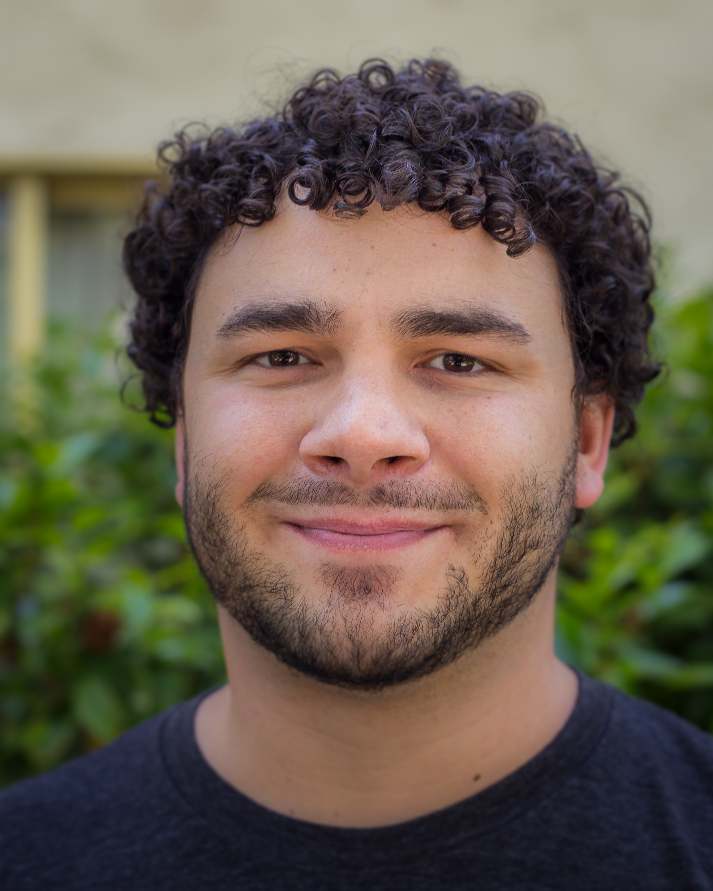 Headshot of Sil smiling. Blurry outdoor background