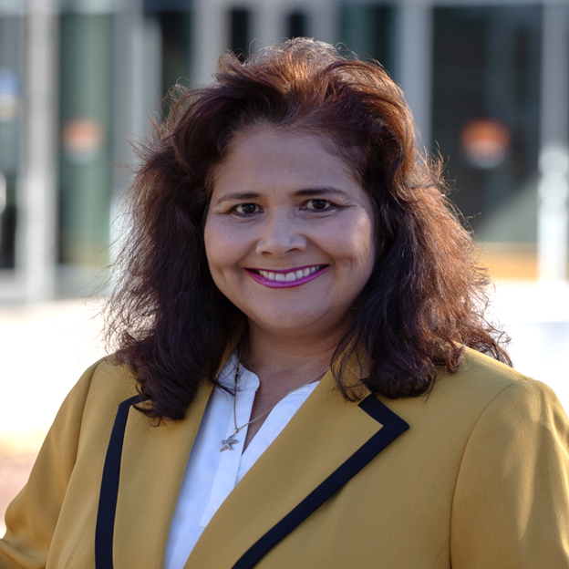 Adela - resized Professional headshot of Adela Cota-Gomez, she smiles wearing a yellow blazer in an outdoor setting.