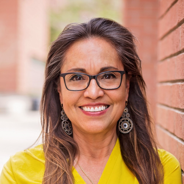 Theresa - resized Professional headshot of Theresa Nino; she leans against a brick wall and smiles in a bright yellow top