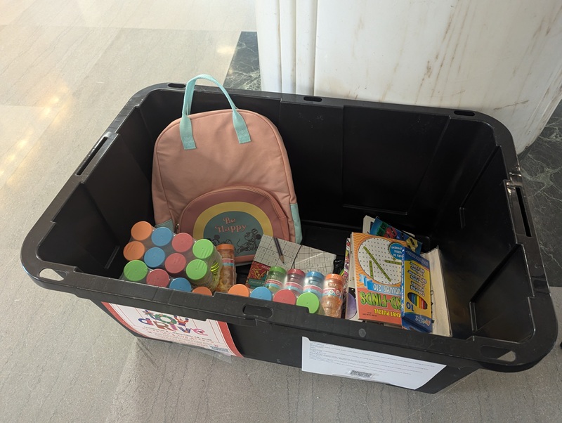 A black storage box with a Toy Drive flier taped to the side contains children's puzzles, blowing bubbles and a backpack.