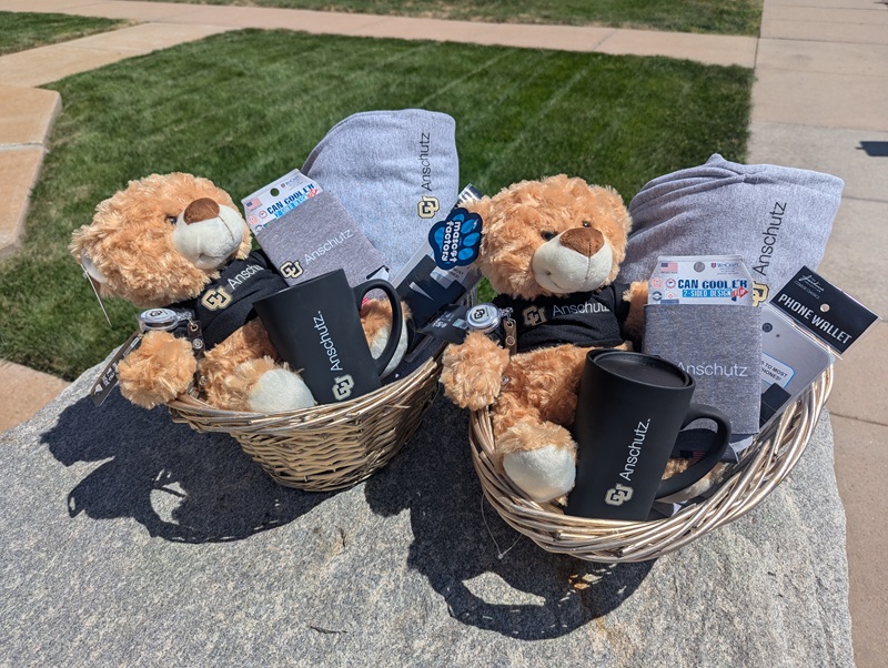 Two golden baskets containing CU Anschutz branded teddy bear, badge holder, koozie, phone wallet, track pants and a mug.