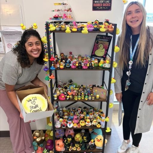 Two people stand on either side of a four-tiered black metal shelf filled with a large collection of colorful rubber ducks. The person on the left holds an open box displaying a round yellow sign that reads 