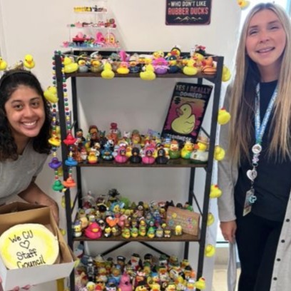 Two people stand on either side of a four-tiered black metal shelf filled with a large collection of colorful rubber ducks. The person on the left holds an open box displaying a round yellow sign that reads 